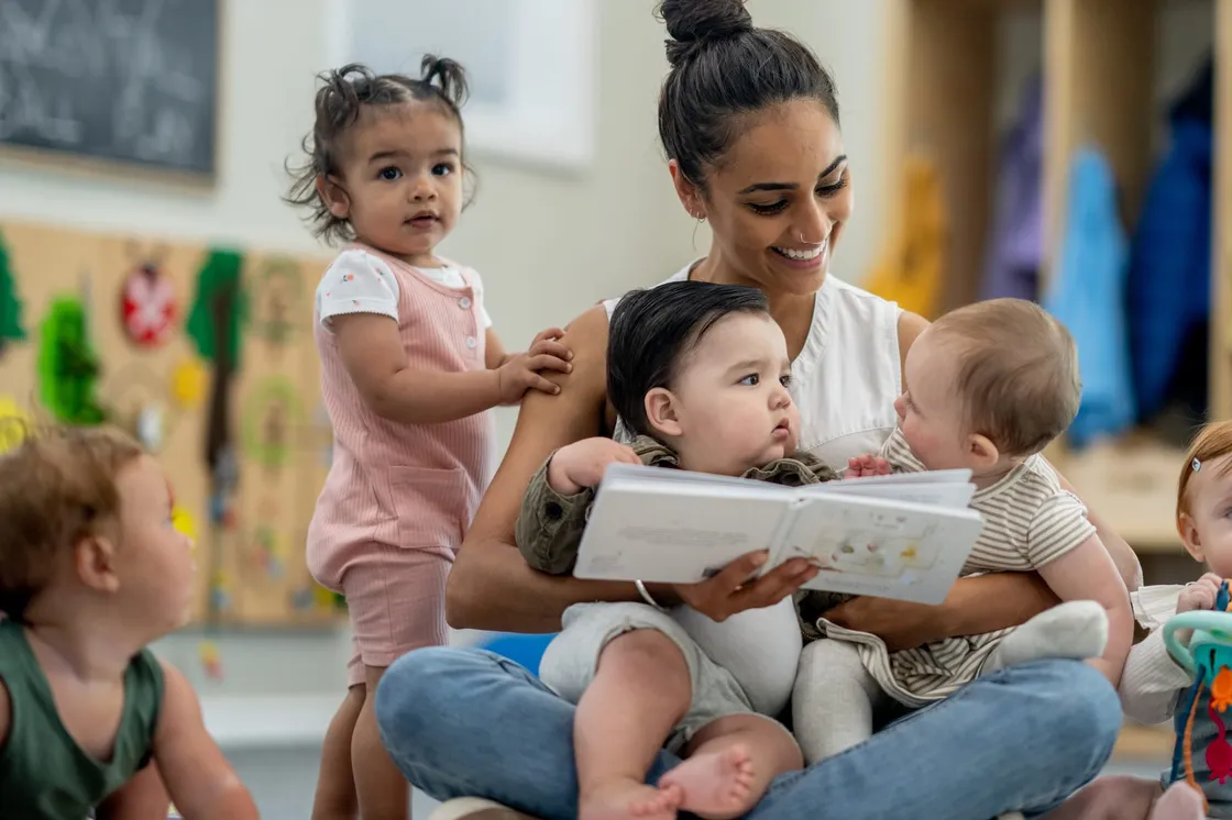 An early childhood educator sitting on the floor of a classroom, reading a picture book to the toddlers and babies gathered in her lap and around her