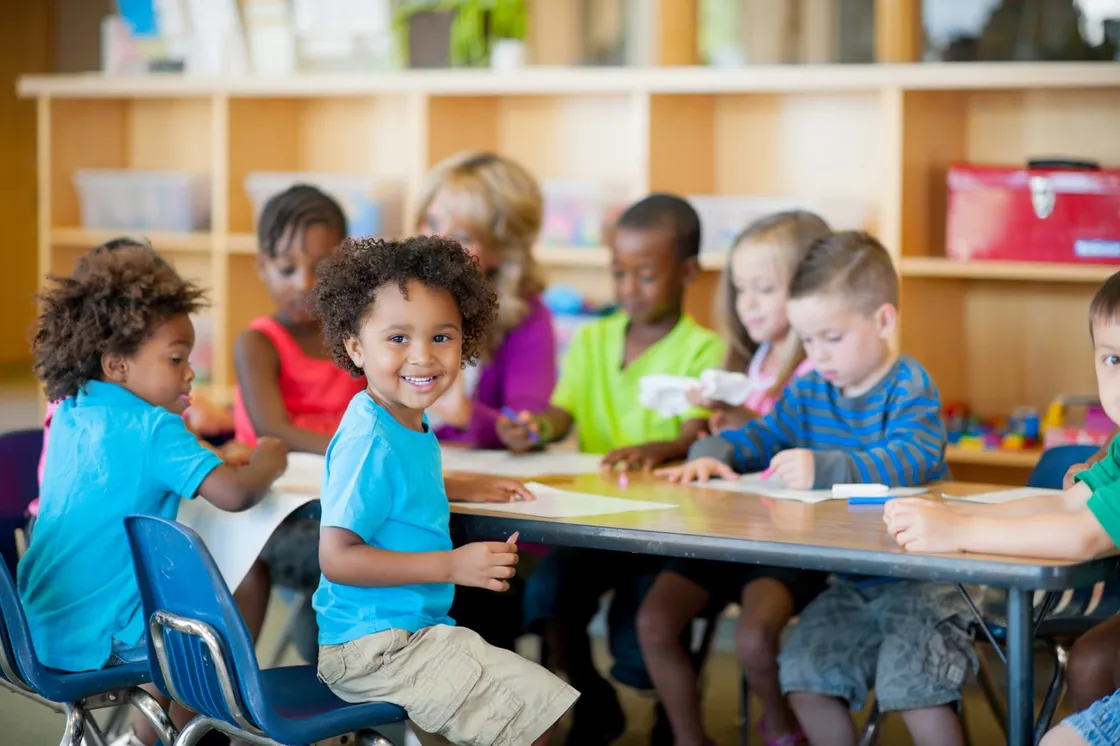 Preschoolers seated around a classroom table with their teacher, one child turning to smile at the camera
