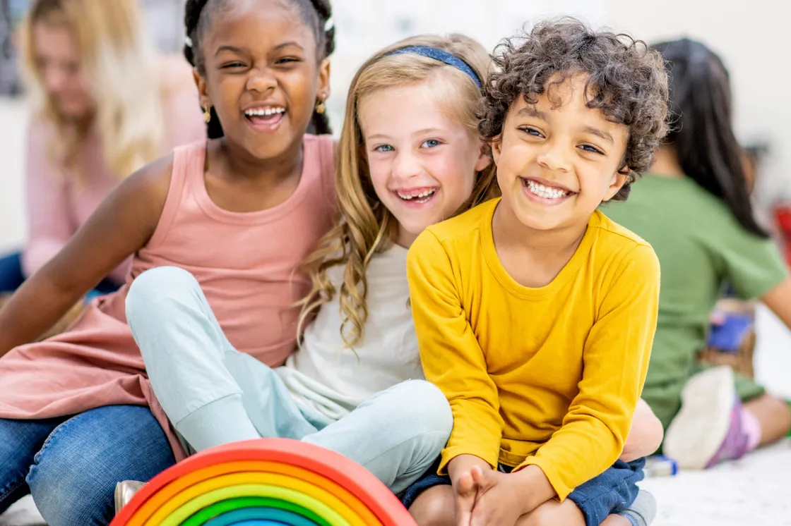 Three young children sitting together on a classroom floor, laughing at the camera beside a wooden rainbow stacking toy