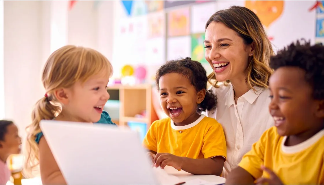 A teacher and three preschoolers laughing together over a laptop in a bright classroom